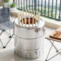 A vertical charcoal grill is shown on a tiled floor of a balcony, with a gray camping chair and a table with skewers.