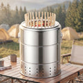 A stainless steel charcoal grill for vertical skewers, sitting on a wooden picnic table with a forest and tents in the background.