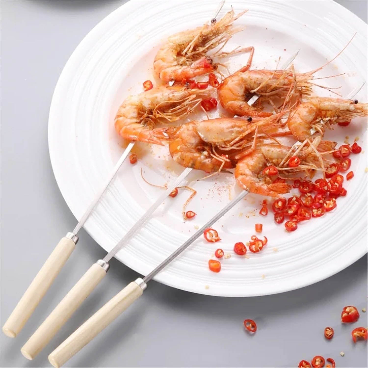 Three metal skewers holding grilled shrimp topped with sliced red chili peppers, presented on a white plate.
