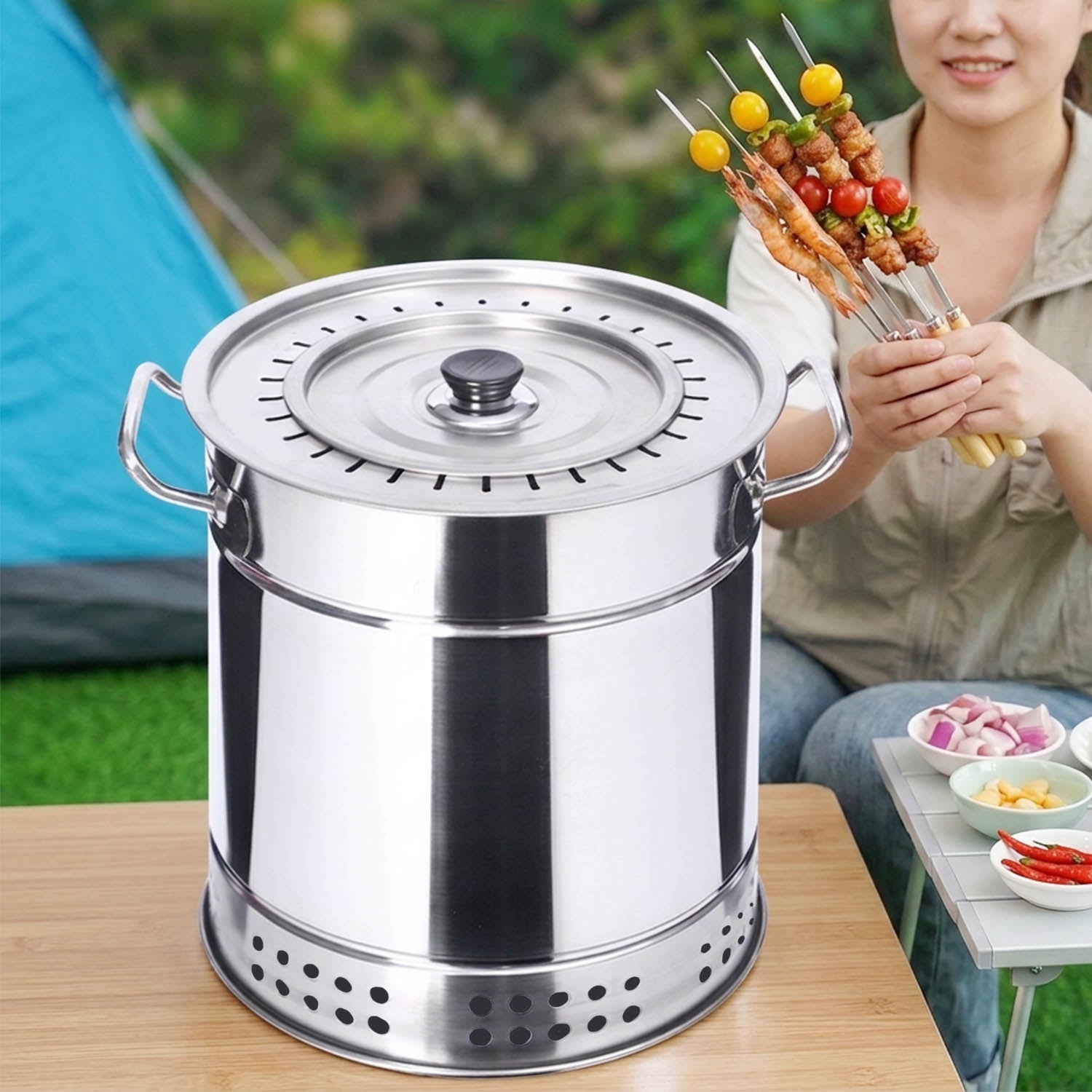A close-up view of a stainless steel vertical charcoal grill, and a woman in the background holding skewers at a campsite.