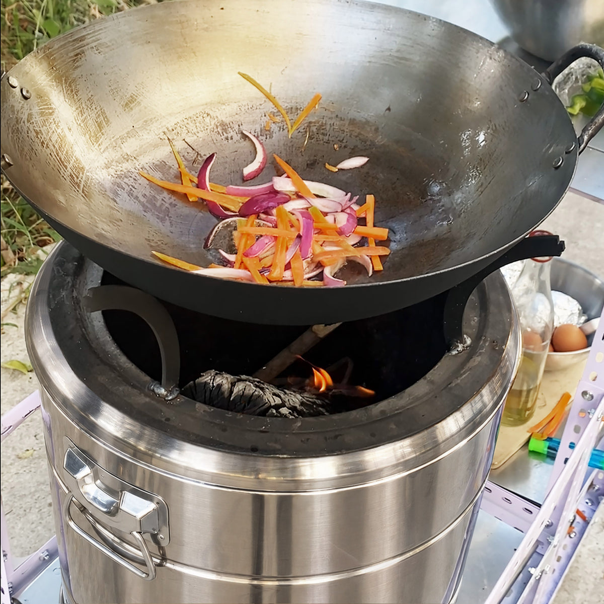 A large stainless steel wok sits on top of a portable outdoor stove with visible flames, cooking sliced onions and carrots. The setup is outdoors on a table with cooking ingredients nearby.