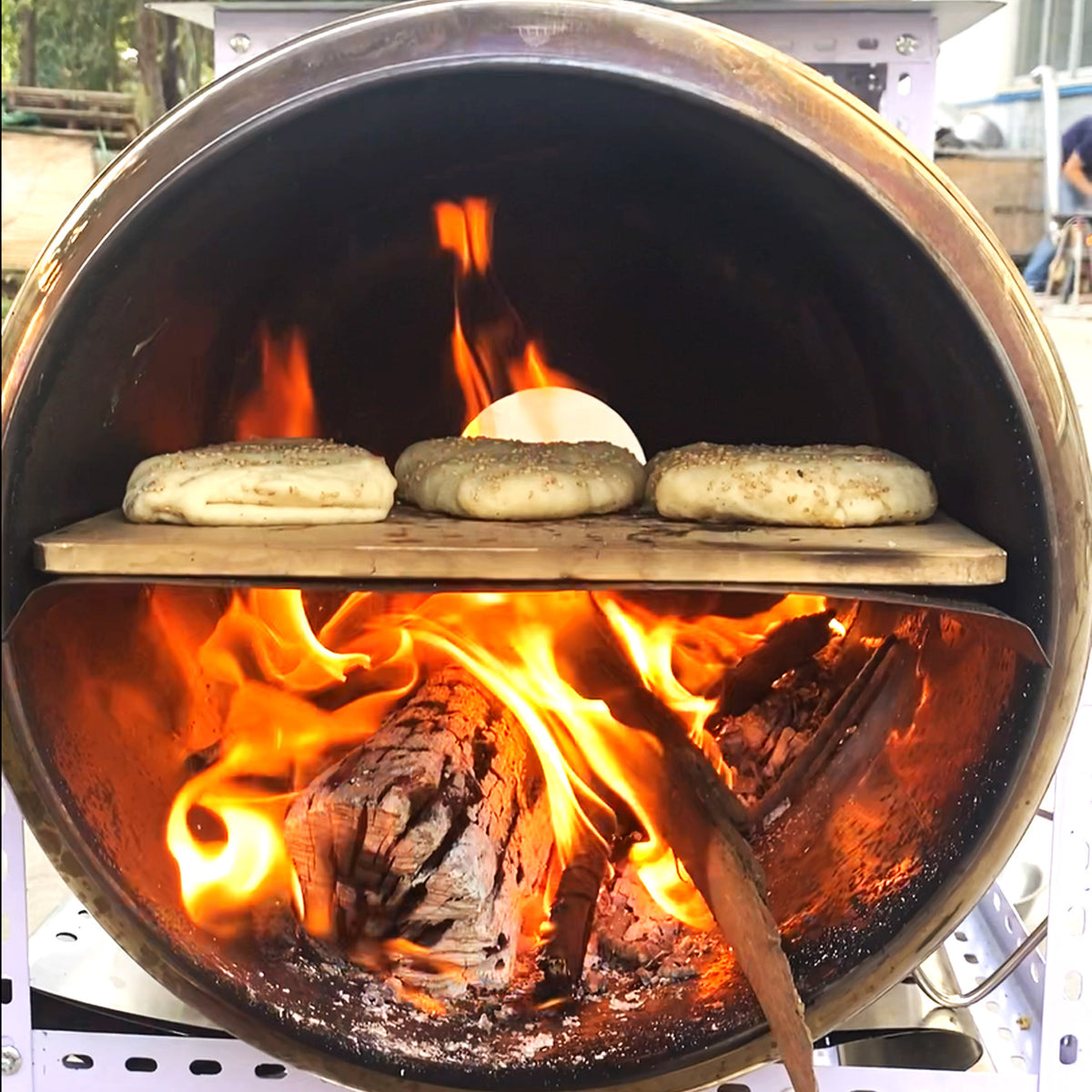 Three sesame-topped flatbreads and a white cheese ball cooking on a wooden shelf inside a large wood-burning oven with bright flames visible.