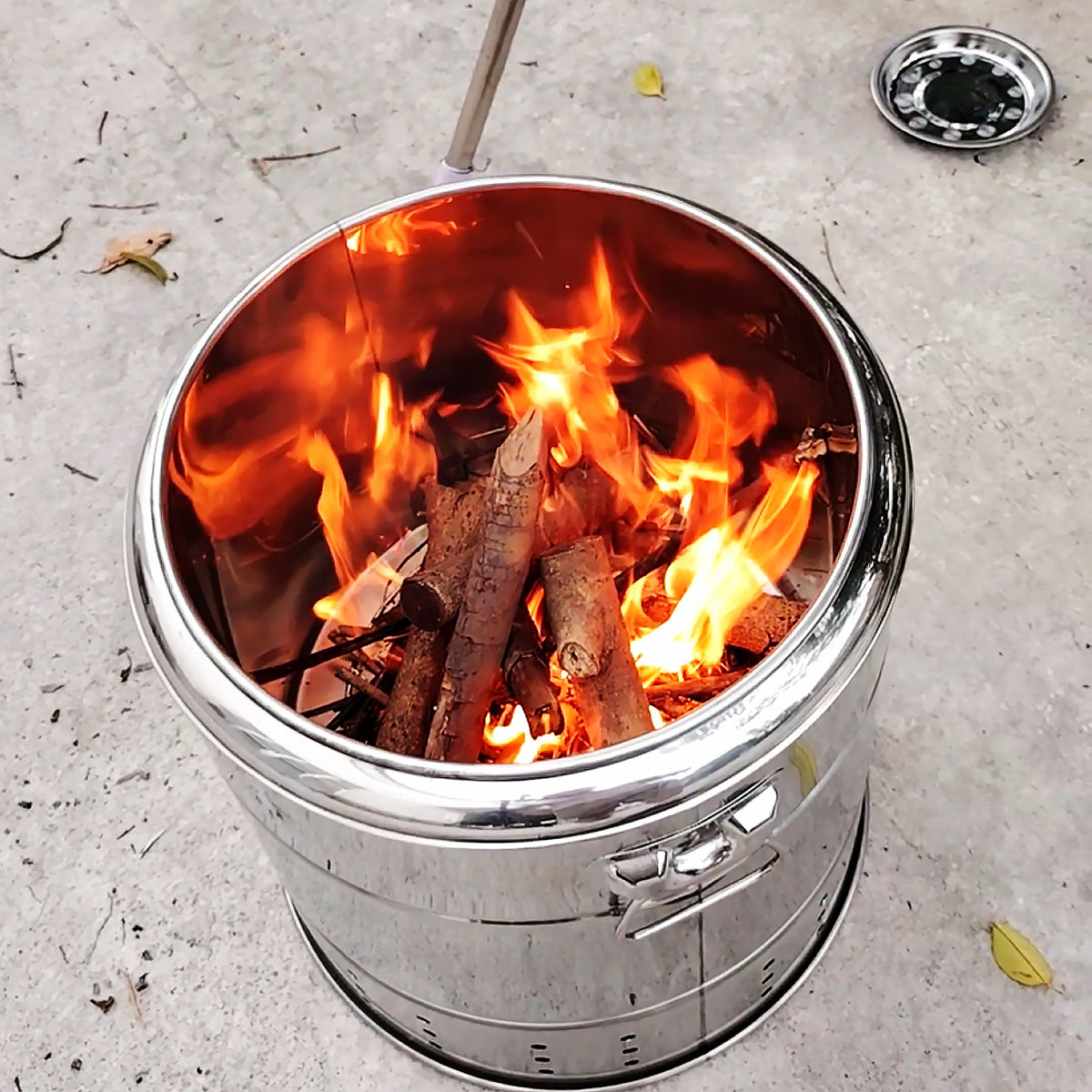 A stainless steel camp stove with a fire burning inside, set on a concrete outdoor floor.