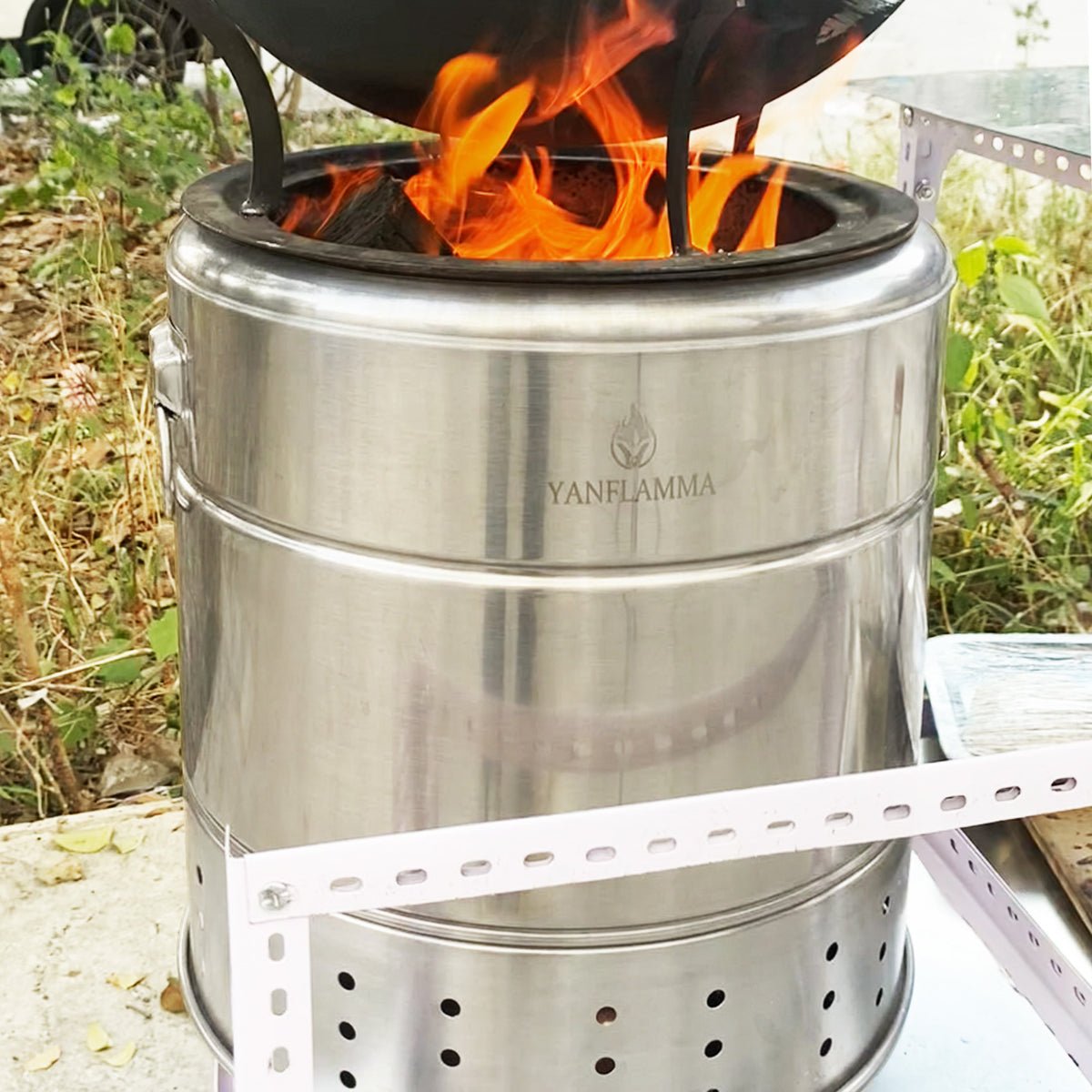 A stainless steel Yanflamma wood stove with visible flames, shown outdoors with grass in the background and a metal support frame in front.